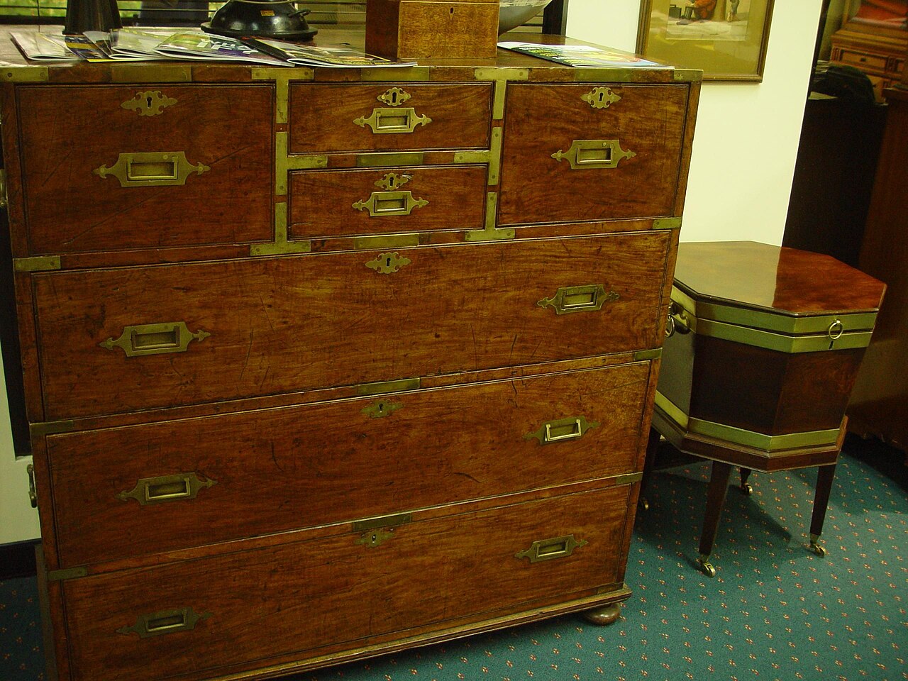 Antique mahogany campaign chest of drawers with brass corners and recessed brass handles, beside an octagonal wine cooler on casters